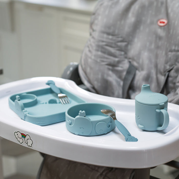 The high chair tray holds the Animal Friends 6-Piece Dinnerware Set - Blue Whale: blue silicone tableware with animal designs, including self-feeding utensils, a cup with lid, bowl, and divided plate. The high chair has a gray cushion.