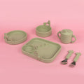 The Nuby Animal Friends 6-Piece Dinnerware Set - Green Giraffe features pastel green silicone baby tableware with giraffe accents, including a handled cup, divided plate, two bowls, spoon, and fork—shown on a pink background.