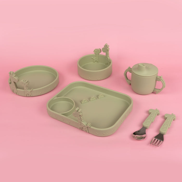 The Nuby Animal Friends 6-Piece Dinnerware Set - Green Giraffe features pastel green silicone baby tableware with giraffe accents, including a handled cup, divided plate, two bowls, spoon, and fork—shown on a pink background.
