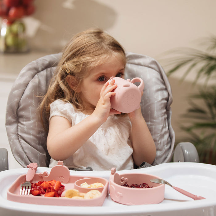 A young child with light brown hair in a white shirt sits in a high chair, self-feeding snacks and fruit from Nuby's Animal Friends 6-Piece Dinnerware Set - Pink Whale, featuring matching divided plate and cup.