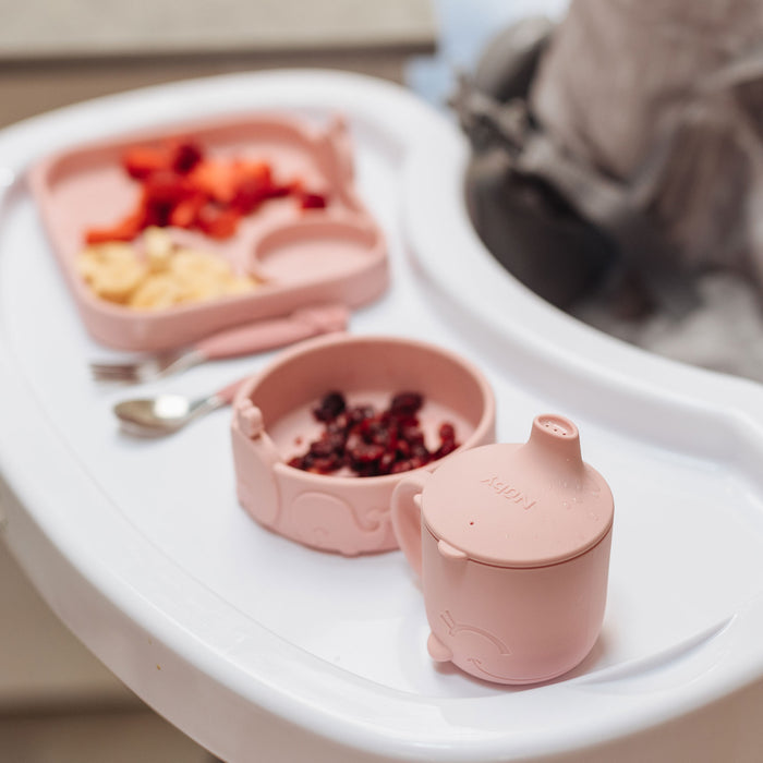 A pink silicone sippy cup, suction bowl with dried berries, and divided plate with banana slices and strawberries are arranged on a white highchair tray, with a fork and spoon nearby.