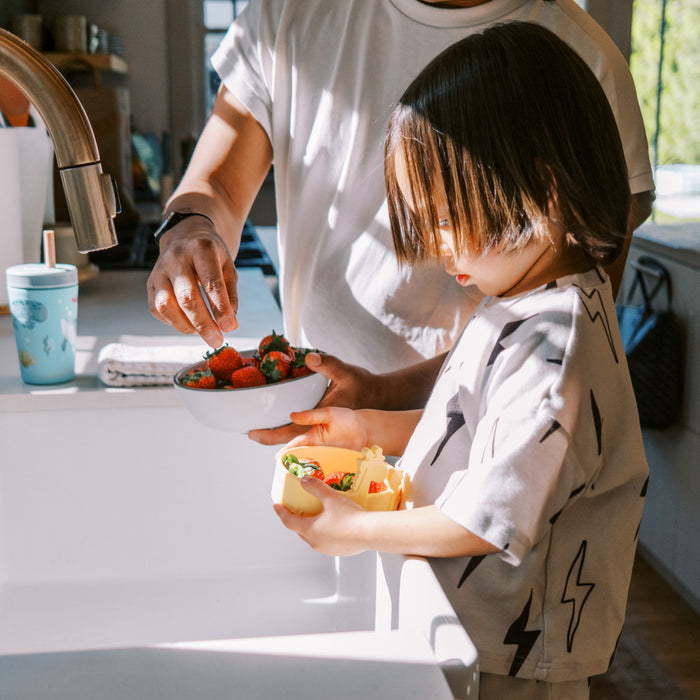 A young child uses the Animal Friends 6-Piece Dinnerware Set - Yellow Giraffe to eat fruit with self-feeding utensils, while an adult reaches for strawberries in a sunny kitchen. A turquoise cup sits by the sink.