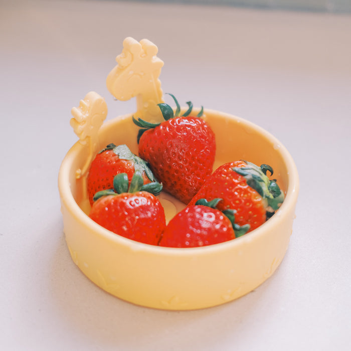 Four fresh strawberries sit in a round, light yellow bowl with two animal-shaped handles from the Animal Friends 6-Piece Dinnerware Set - Yellow Giraffe, placed on a light-colored surface.