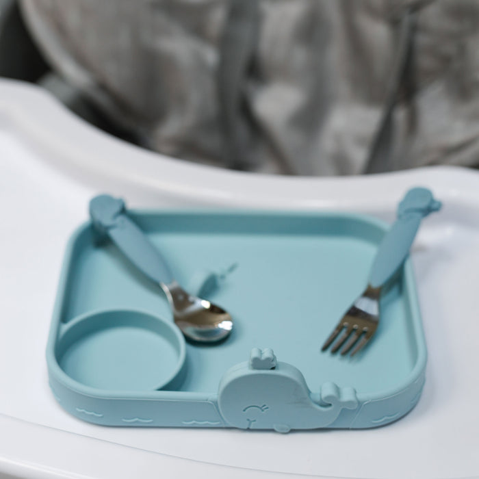 The Nuby Animal Friends All Silicone Tray, a blue whale-shaped toddler plate with a round compartment, sits on a white highchair tray. The matching spoon and fork feature whale designs on their handles.