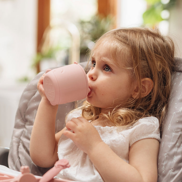 A young child with blonde hair sits in a high chair, wearing a white top and sipping from a pink cup while looking slightly upward. The softly blurred background features green plants, creating a calm, nurturing atmosphere.