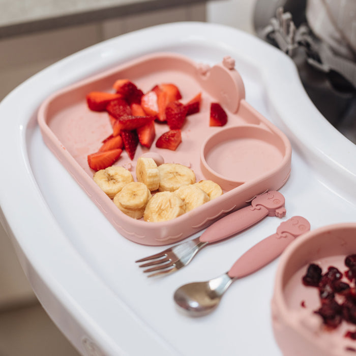 A pink baby plate with strawberries and bananas sits on a high chair tray beside the Nuby Animal Friends Silicone & Stainless Steel Utensils and a small bowl of fruit—perfect for little ones learning self-feeding.