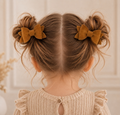 Child with brown hair styled in buns with velvet bows, wearing a beige knit sweater.