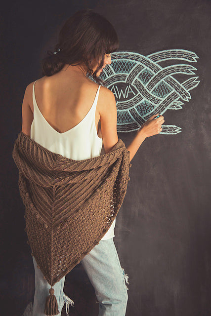 Image of back of woman wearing white tank top and brown shawl drawing on chalkboard the word "AWA" inside intricate pattern