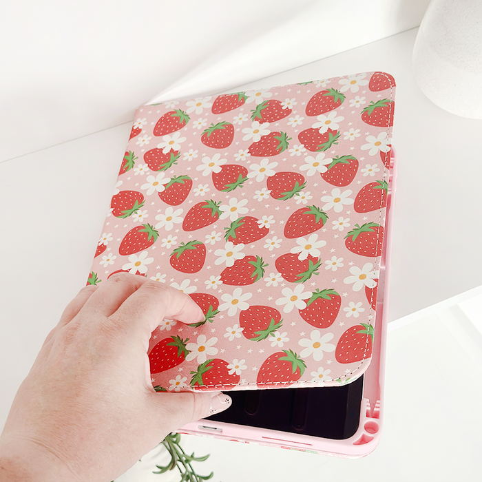 Hand opening Coconut Lane pink iPad case with strawberry and daisy pattern, vegan leather, on white table.