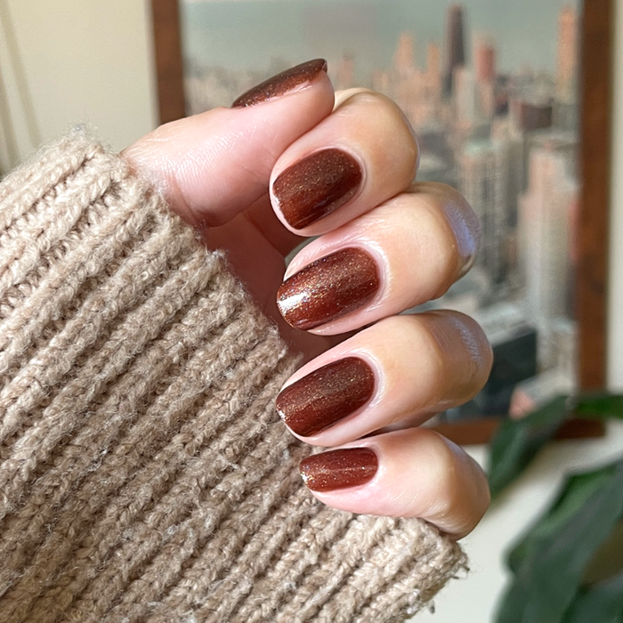 Hand with short nails painted in deep shimmery brown Museum Date gel polish wearing beige knit sleeve indoors