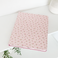 A pink notebook adorned with a delicate floral pattern rests on a white surface. A plant with lush green leaves is partially visible in the corner, and to the right, a white cylindrical object, likely a vase, is seen in the background. The setting perfectly complements my Coconut Lane Ditsy Floral Pink iPad Case.