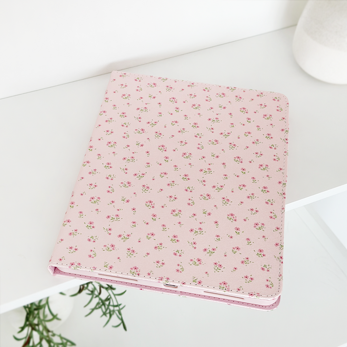 A pink notebook adorned with a delicate floral pattern rests on a white surface. A plant with lush green leaves is partially visible in the corner, and to the right, a white cylindrical object, likely a vase, is seen in the background. The setting perfectly complements my Coconut Lane Ditsy Floral Pink iPad Case.