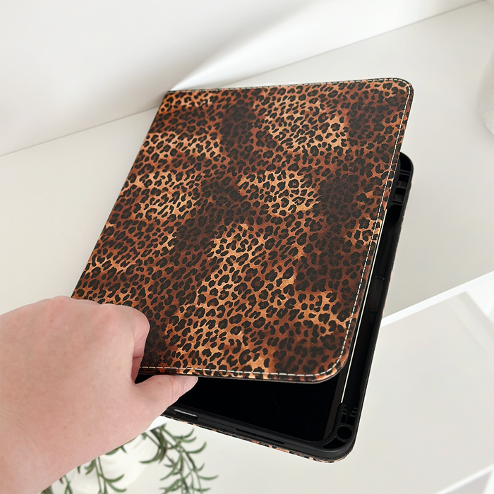 A hand closes a Coconut Lane iPad case with a leopard print on a white surface, next to a green plant.