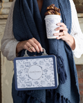 Person holding a blue-and-white floral tin lunch box with the “Maman” logo and a silver handle.