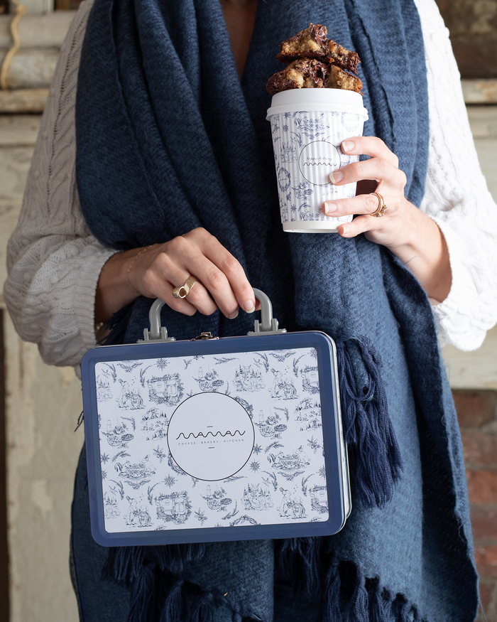 Person holding a blue-and-white floral tin lunch box with the “Maman” logo and a silver handle.
