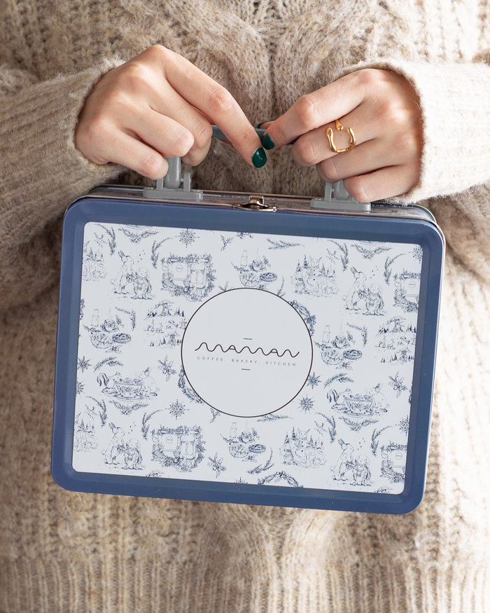 Person holding a blue-and-white floral tin lunch box with the “Maman” logo and a silver handle.