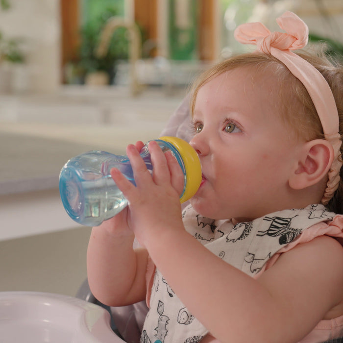 A young child in a high chair drinks from a blue and yellow Nuby Mini Gripper Soft Spout Sippy Cup (2 Pack), wearing a bow headband and bib in a bright kitchen.