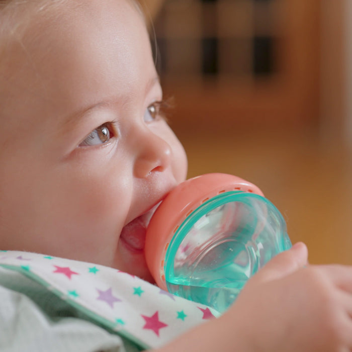 A smiling baby wears a star-patterned bib and drinks from the Nuby Mini Gripper Soft Spout Sippy Cup (2 Pack) with a teal body and orange lid, perfect for little hands.