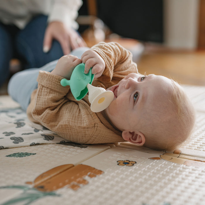 A baby lies on a patterned playmat, chewing on the green and white Nuby Mushroom Bobble Head Teether Toy while looking up. An adult’s hand appears in the background.