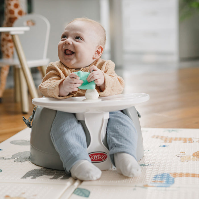 A smiling baby in a beige sweater and light blue pants sits on a gray Nuby seat, holding the Nuby Mushroom Bobble Head Teether Toy. The baby is on a play mat with a chair, table, and blurred home interior in the background.