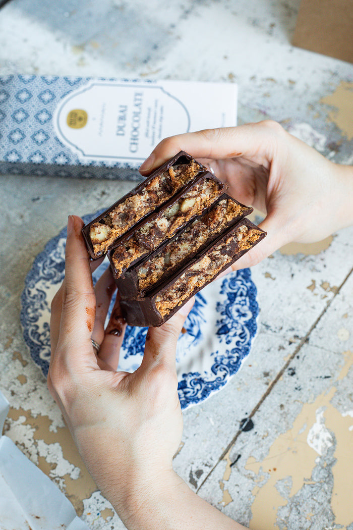 Hands holding a cross-section of a dubai chocolate bar, with a blue-and-white patterned plate and a box labeled 'DUBAI CHOCOLATE' in the background.