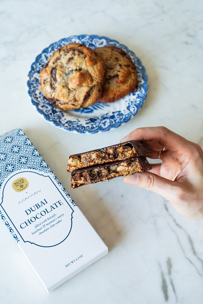 A hand holds a chocolate-dusted cookie bar in half, with two cookies on a blue-and-white patterned plate in the background and a white box labeled 'Dubai Chocolate' on a light marble surface.