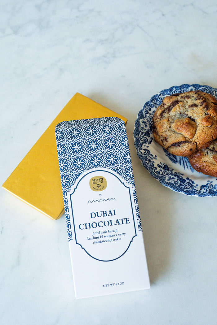 A package of Nuts Factory Dubai Chocolate cookies lies on a white marble surface next to a yellow notebook and a blue-and-white patterned plate with chocolate chip cookies.