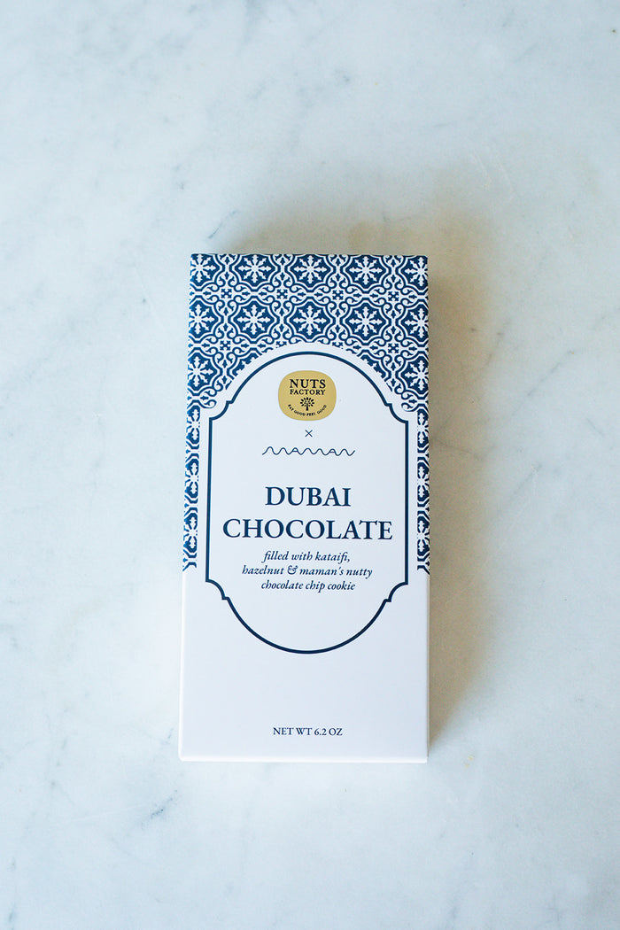 A boxed bar of Nuts Factory Dubai Chocolate, featuring a white box with a blue geometric pattern border and a gold logo seal, set on a light marble surface.