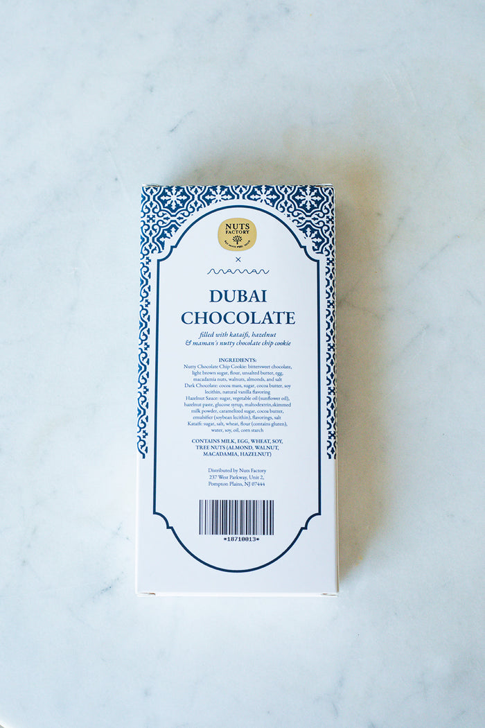 A rectangular box of Nut Factory Dubai Chocolate, featuring a white background with a blue ornate border and black text, photographed flat on a light marble surface.