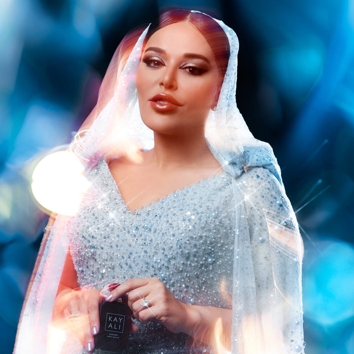 Mona Kattan in a sparkling outfit holding a product with 'KAYALI' branding against a blue blurred background