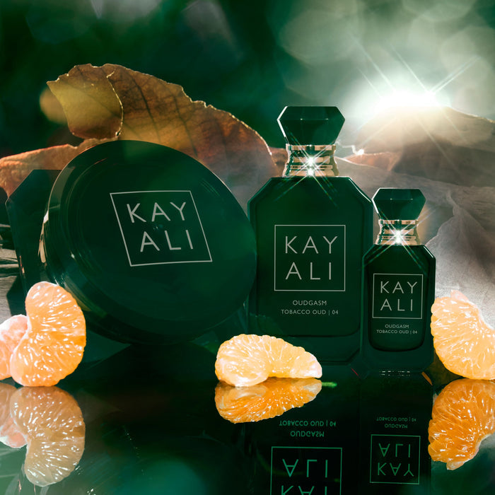 Two green perfume bottles with 'KAYALI' branding surrounded by orange slices and leaves on a reflective surface.