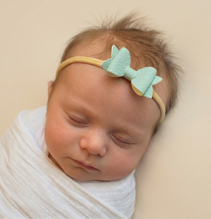 Newborn baby sleeping, wearing a mint green bow headband on a beige background