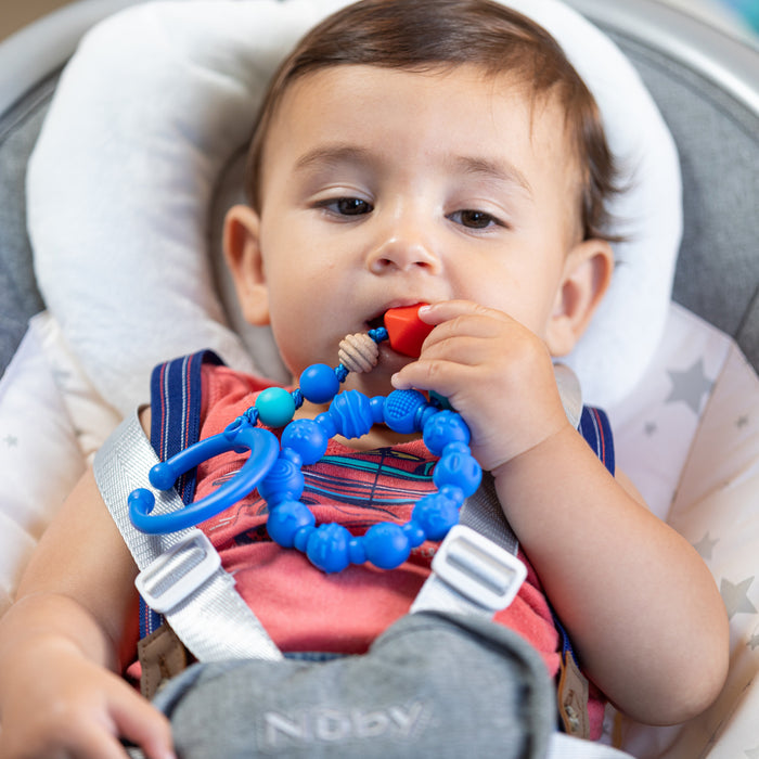 A baby in a car seat, wearing a red shirt and grey straps, chews on the blue Nuby Tag-a-Long Teether, which stands out against the softly blurred background.