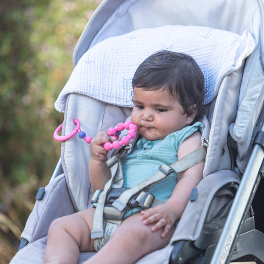 A baby with dark hair sits in a stroller, holding the Nuby Tag-a-Long Teether (pink) and wearing a light blue outfit. A white pillow supports the baby's head.