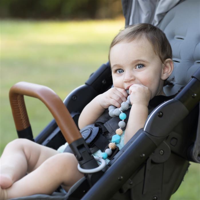 A blue-eyed baby smiles in a stroller outdoors, holding the colorful Nuby Tag-a-Long Teether and looking at the camera.