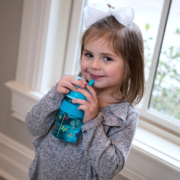 A young girl with a white bow smiles while drinking from a Nuby Thirsty Kids FREE STYLE Hard Straw Water Bottle, standing by a window in a bright room.