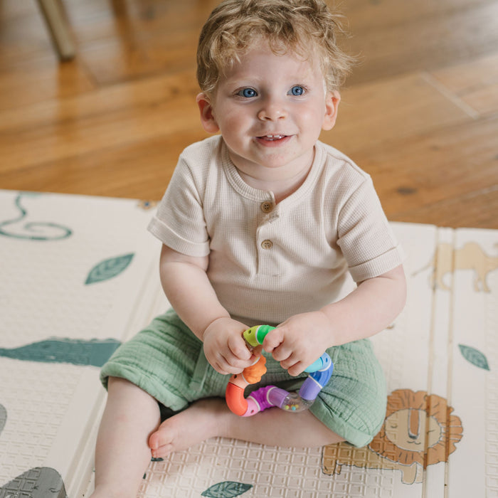 A smiling toddler in a beige shirt and green pants sits barefoot on a wooden floor, holding the Twista Rattle Teether Toy | Rattle Balls on an animal and leaf-themed play mat with a lion, perfect for teething relief.