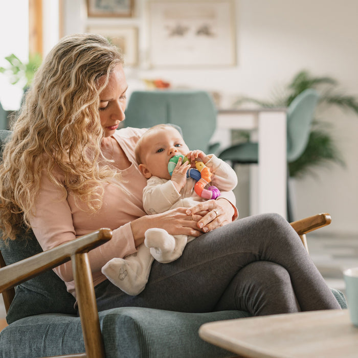 A woman with long curly hair sits in a cozy living room, holding an infant in her lap. The baby, gazing forward, plays with a colorful teething toy under the warm, soft lighting.