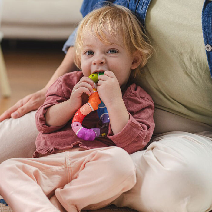 A blonde-haired toddler in a pinkish shirt and light pants sits on an adult’s lap, curiously looking at the camera while chewing on and enjoying the colorful Twista Rattle Teether Toy | Rattle Balls.