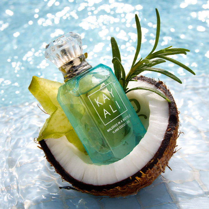 Green perfume bottle labeled 'KAYALI' on a split coconut with a leaf against a sparkling water background.