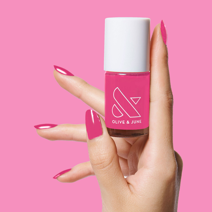 Hand holding Olive and June WILD ORCHID polish bottle with matching smooth, glossy magenta manicure on pink background