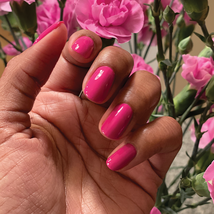 Hand with glossy WILD ORCHID nail polish in vibrant magenta purple against a backdrop of pink flowers